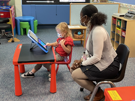 Young female student works on an iPad while a female paraeducator sits nearby.