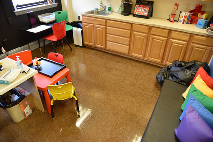 Room with furniture in bright primary colors, brown laminate flooring, and wooden cabinets. There's a toy crab on top of the main red kid's table on a black tray. Pillows in the color of the rainbow are against the wall on top of a mat.