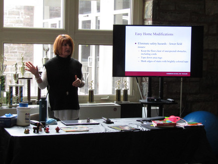 Woman standing in front of table giving a presentation. To the right is a TV displaying a slide from a presentation.